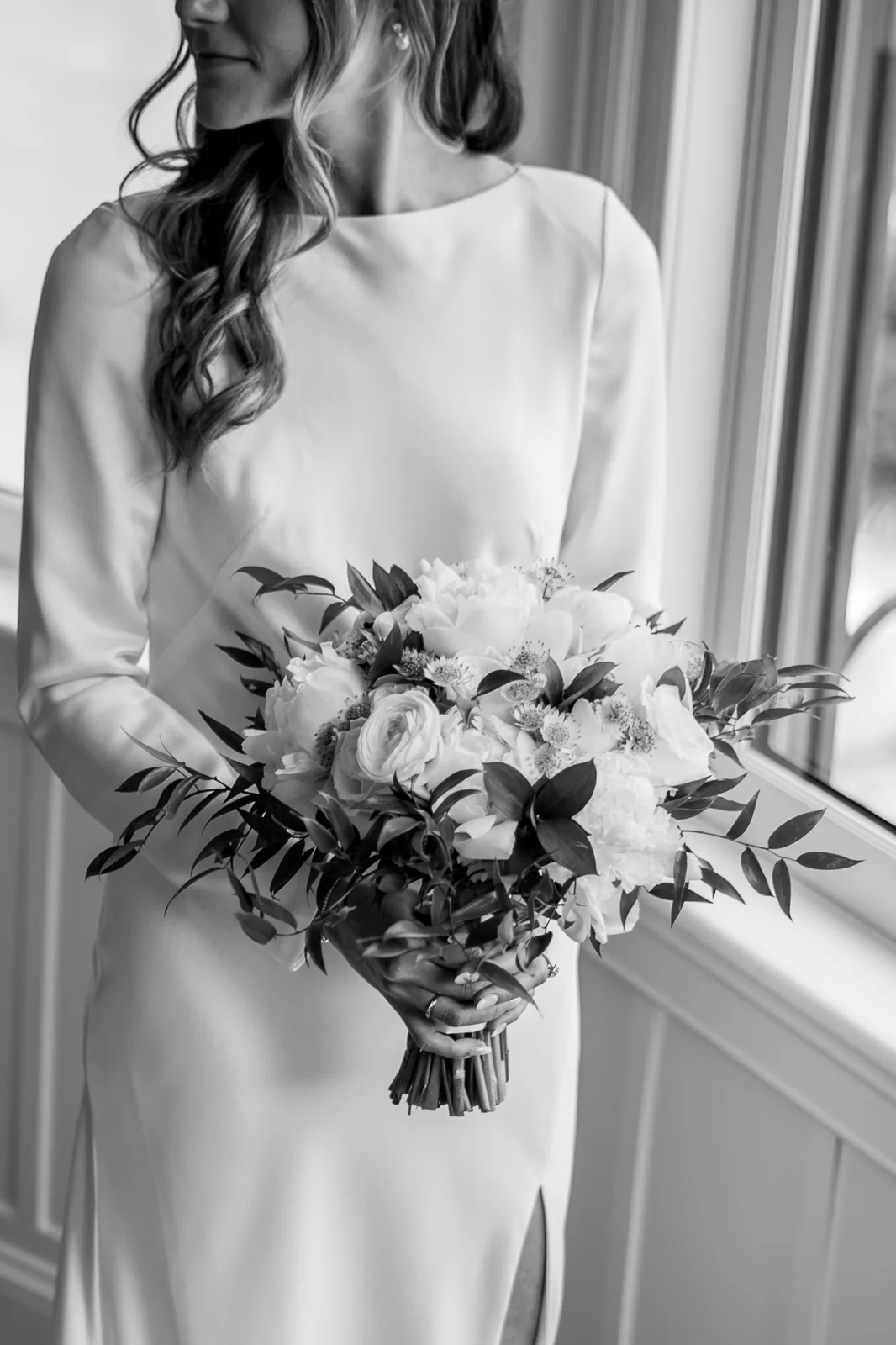 Black and white detail of the bride holding a peony and ranunculus bouquet in window light