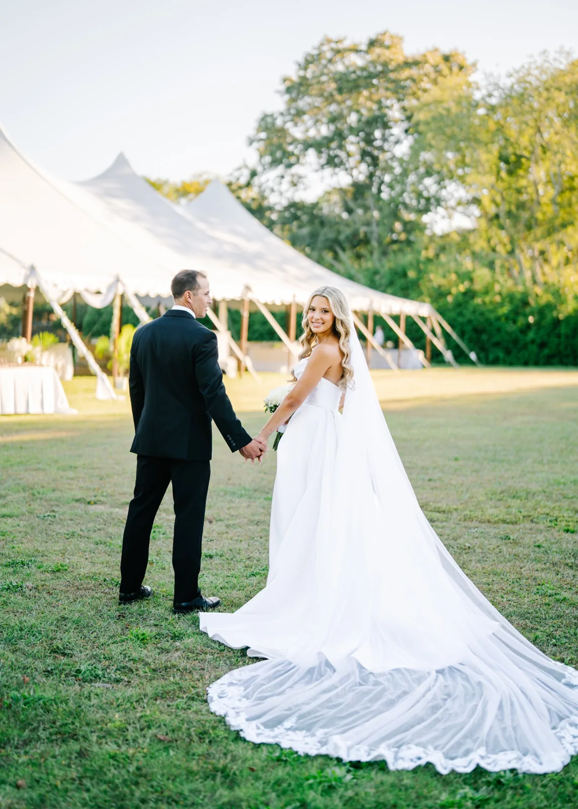 Bride glances back walking hand in hand toward a sailcloth tent, Hamptons wedding photography