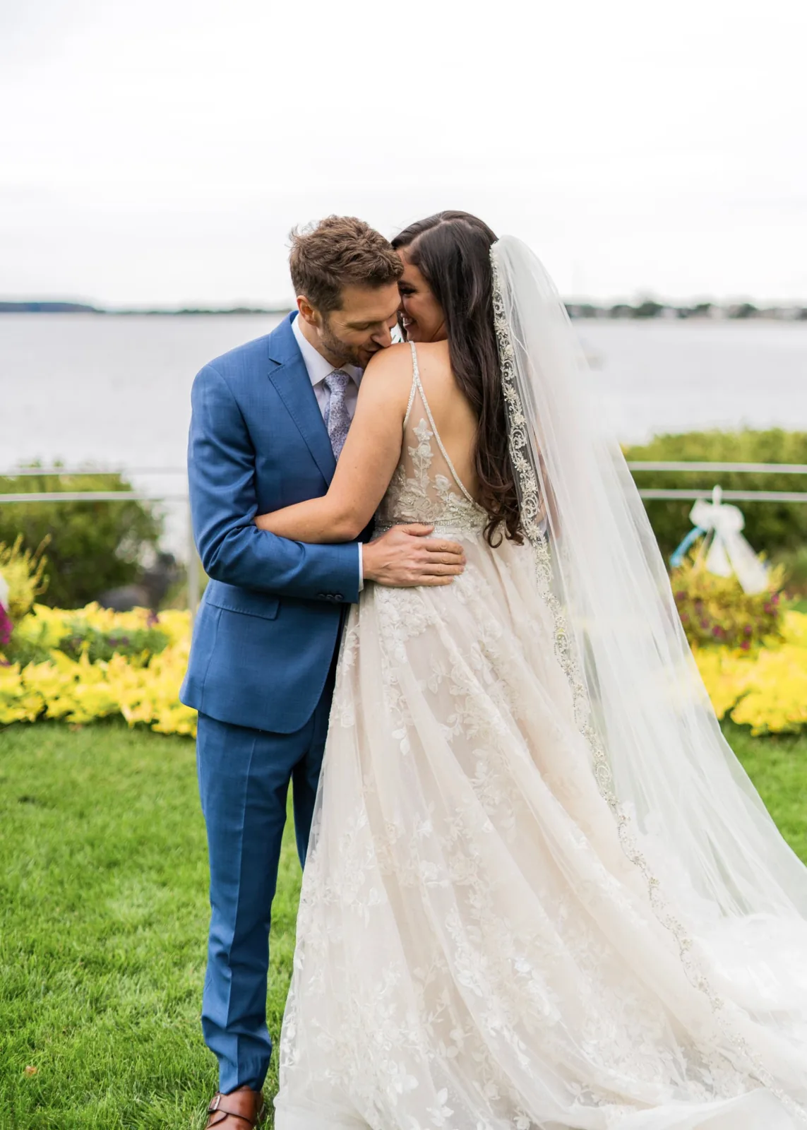 Groom kisses the bride's shoulder beside the water, lace gown and beaded veil, Long Island waterfront wedding